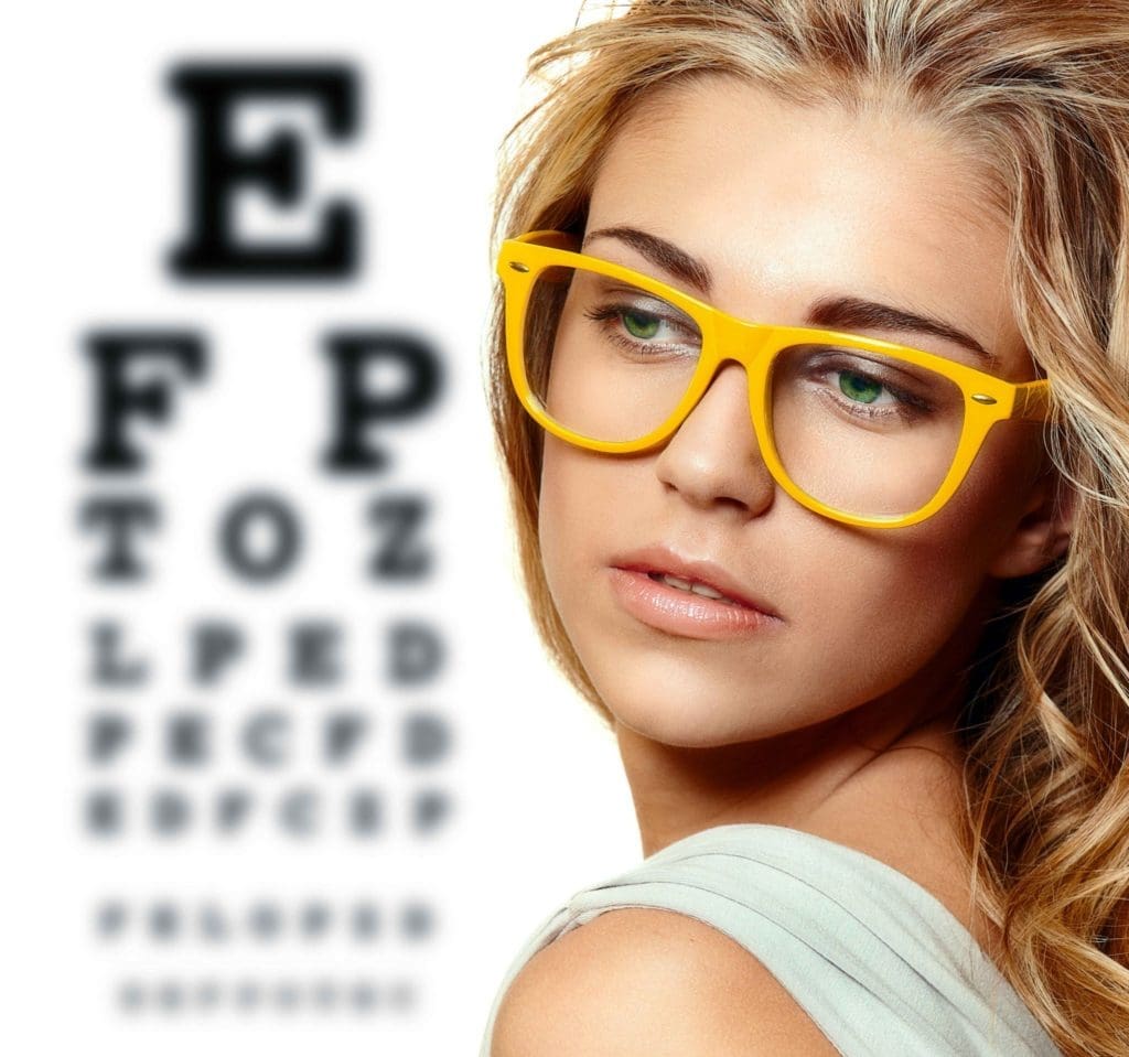 A young woman wearing glasses stands in front of an eye chart, highlighting the importance of detecting eye problems early with optometrists.