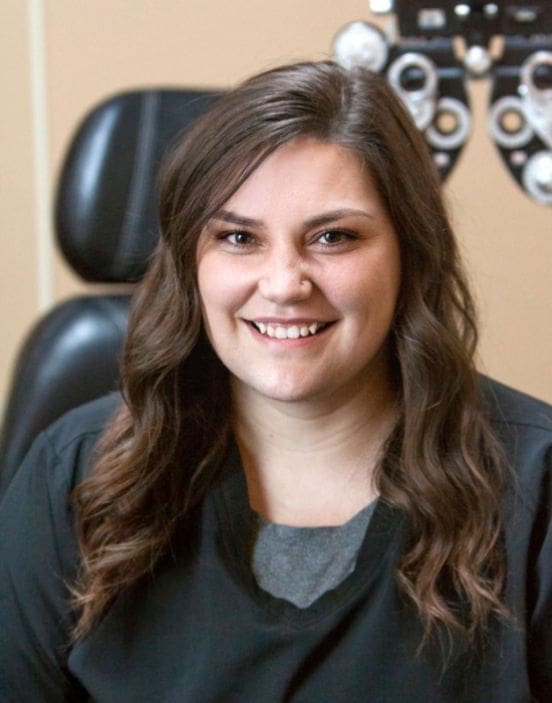 A smiling staff member with long hair sits in an office chair in Idaho Falls, with optometric equipment in the background.