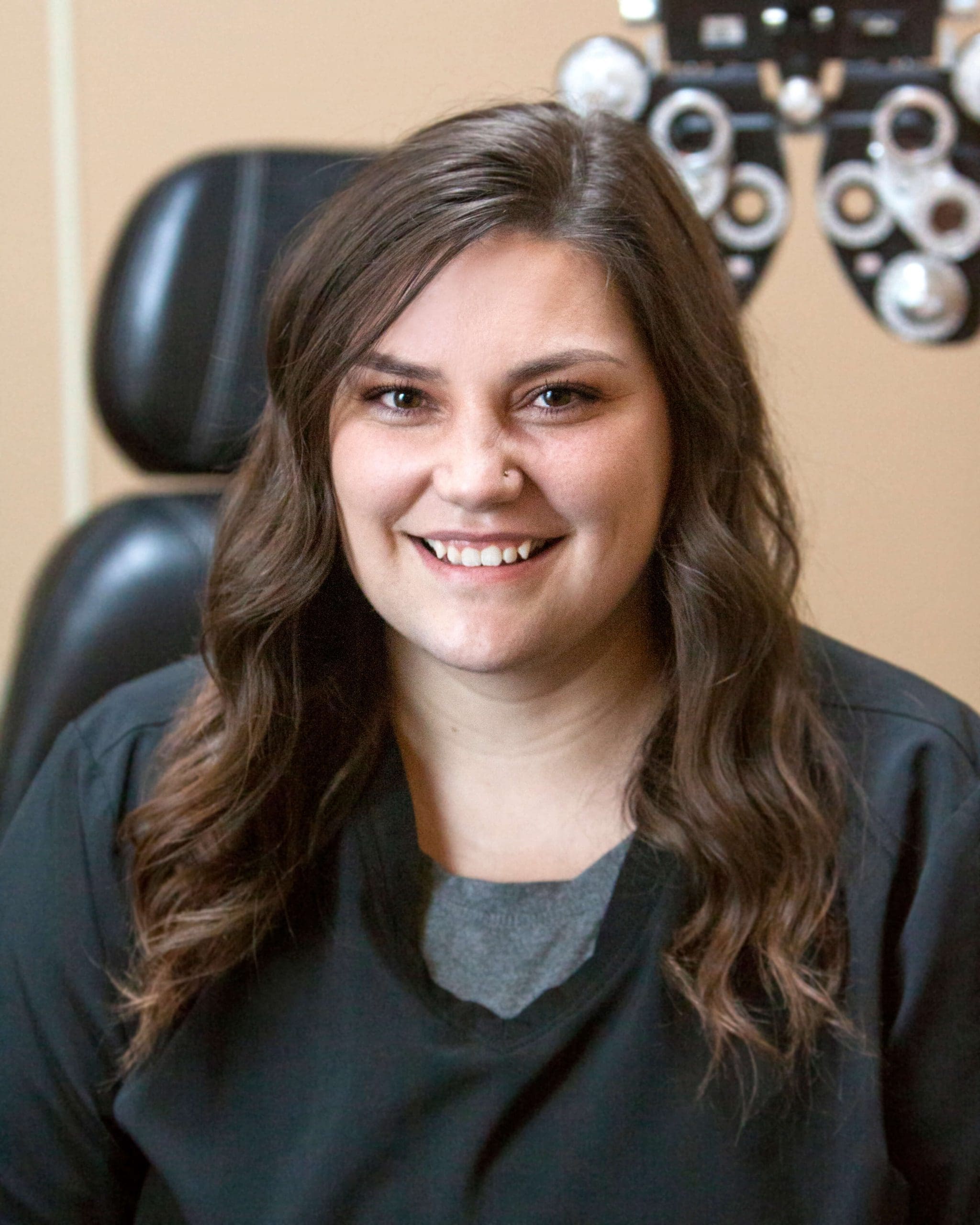 A smiling woman in a black shirt sitting in front of an eye chart at an Eye Doctor Idaho Falls office.