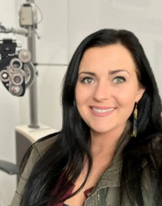 Woman with long dark hair smiling in front of optometry equipment, specializing in administrative support.