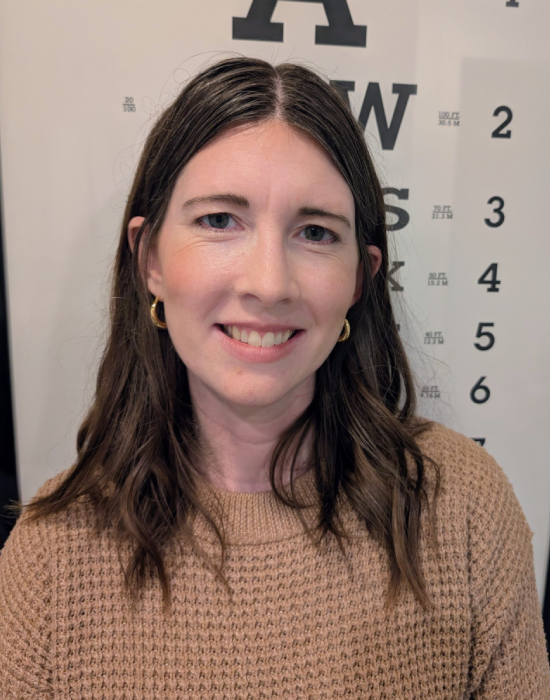 A person with long brown hair and gold hoop earrings smiles warmly, dressed in a brown sweater. Behind them, an eye chart hints at their focus on providing top-notch administrative support.