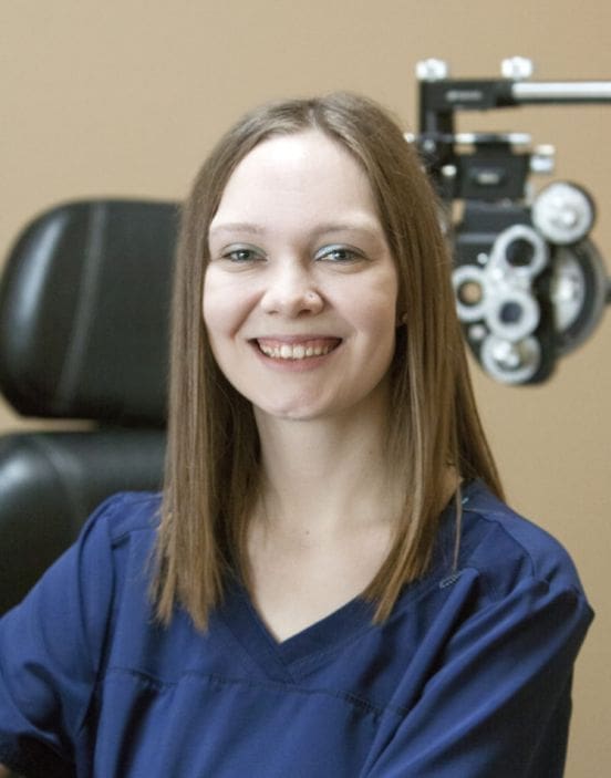 A smiling staff member in medical scrubs with an ophthalmology phoropter in the background in Idaho Falls.