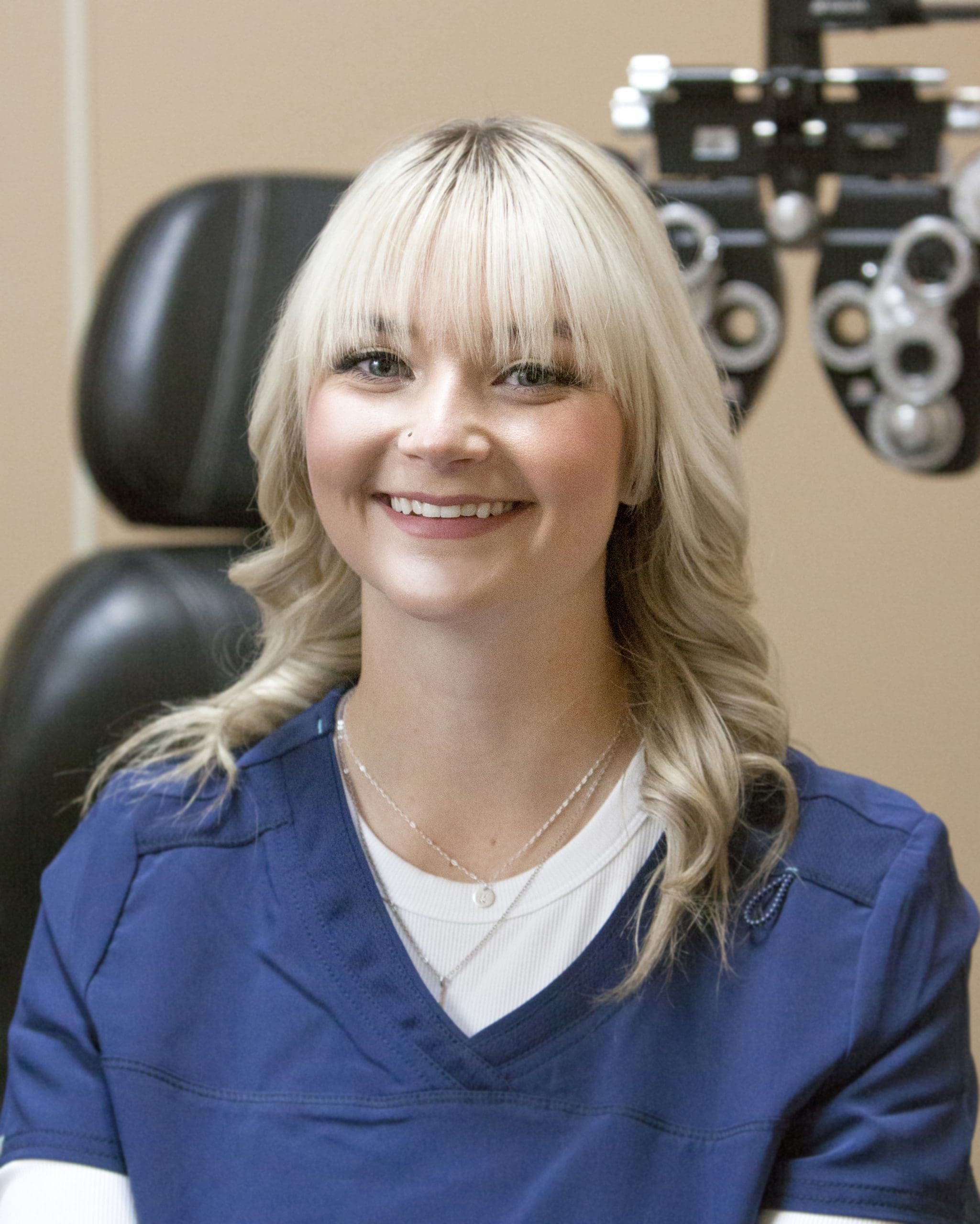 A smiling woman in a blue scrub sitting in front of an eye exam at Eye Doctor Idaho Falls.