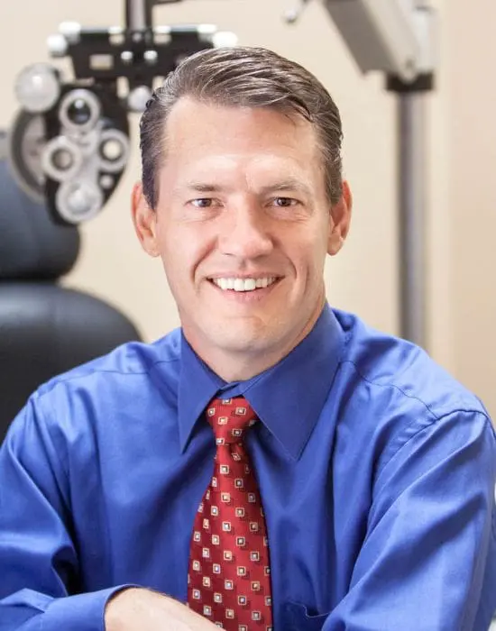 A smiling member of the management team in professional attire with an ophthalmologist's phoropter in the background.