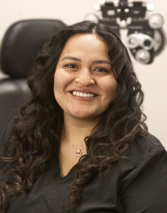 A smiling woman with long curly hair, wearing a black outfit and a necklace, seated in front of an office chair at the Twin Falls staff office.