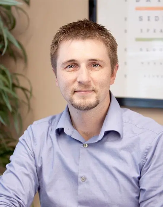 A professional headshot of a smiling man with light stubble, wearing a blue button-up shirt, seated indoors, from the management team.