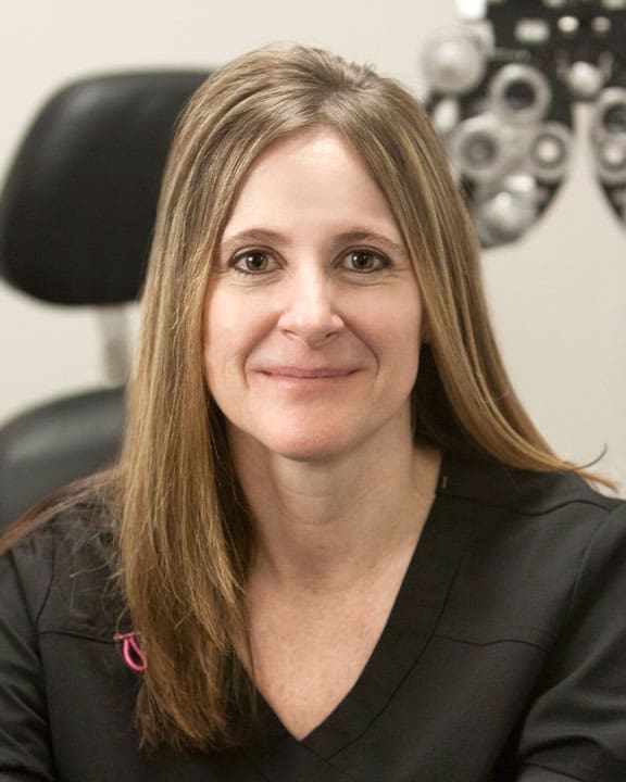 A woman in a black shirt smiling in front of a pair of eye glasses at an Optometrist Ogden office.