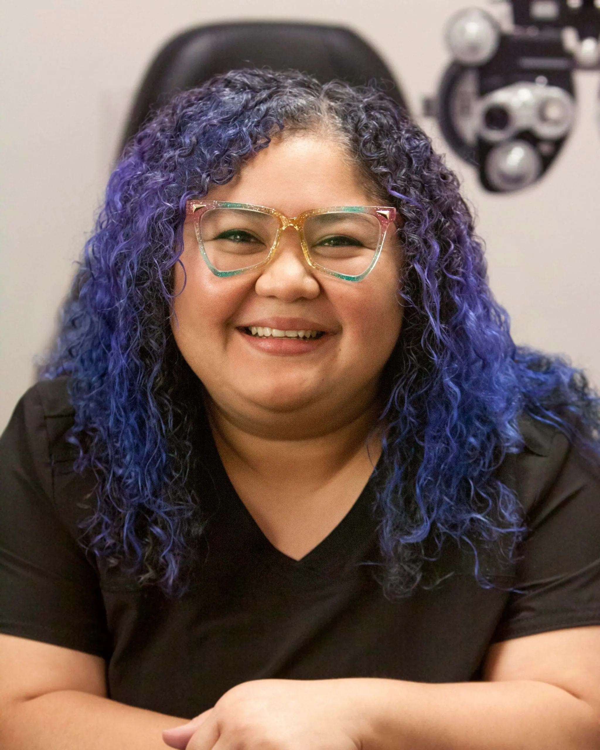 A smiling woman with blue curly hair in front of a pair of glasses at an eye doctor in Twin Falls.