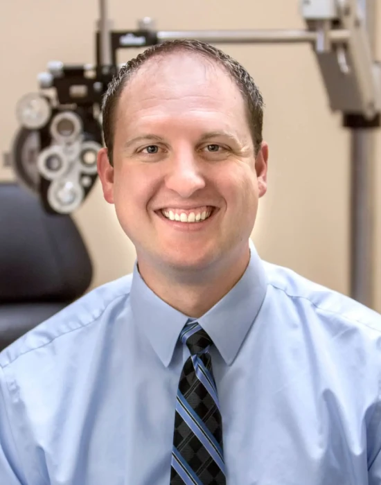 A man in a light blue dress shirt and striped tie, part of the management team, smiles while seated in an optometrist's office, with eye examination equipment in the background.