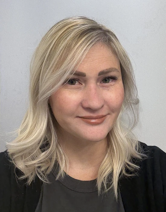 A woman with shoulder-length blonde hair and light makeup smiles at the camera against a plain light background.