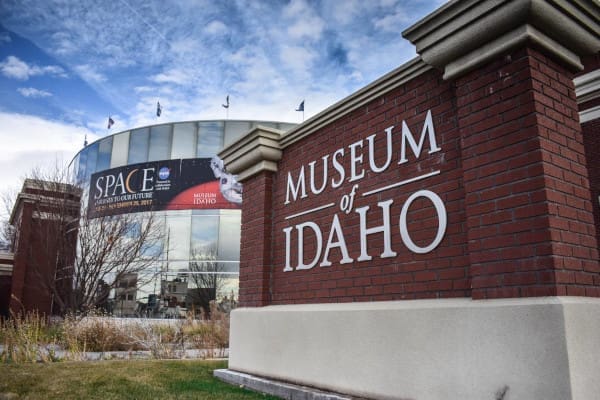 The museum of Idaho sign, showcasing various locations, is in front of a building.