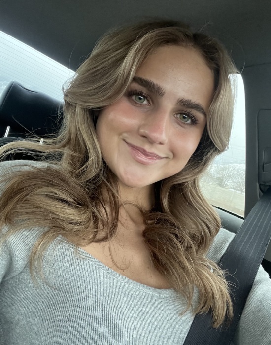 A young woman with long hair, wearing a gray top and a seatbelt, smiles while sitting inside a car, perhaps on her way to provide exceptional Patient Services.