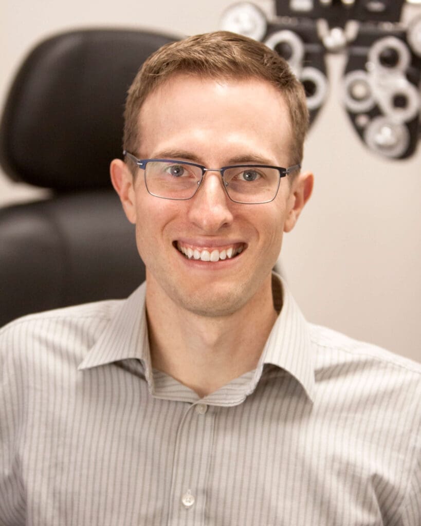 A man in glasses is smiling in front of an eye chart at an optometrist in Idaho Falls.