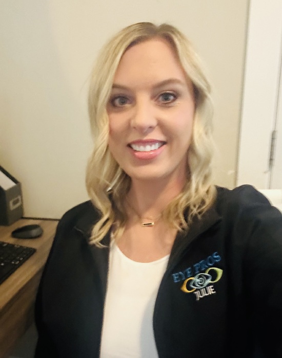 A woman with blonde hair is smiling at the camera. She is wearing a black jacket with an "Eye Pros" logo and sitting at a desk, providing patient services. A computer keyboard is partially visible beside her.