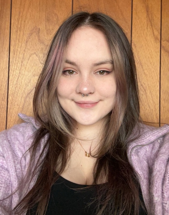 A young person with long hair and a nose ring smiles confidently at the camera, wearing a purple jacket and a black top, standing in front of a wooden background, embodying a patient demeanor.