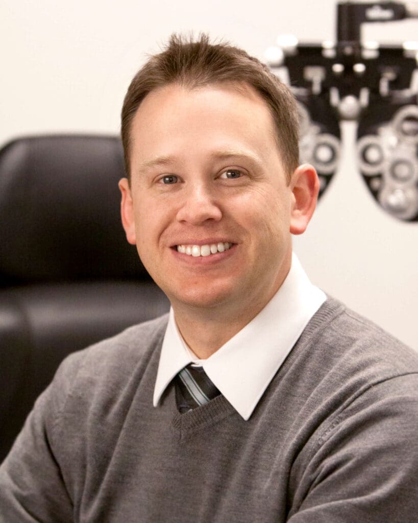 A man in a gray sweater sitting in front of an eye chart meets doctors.