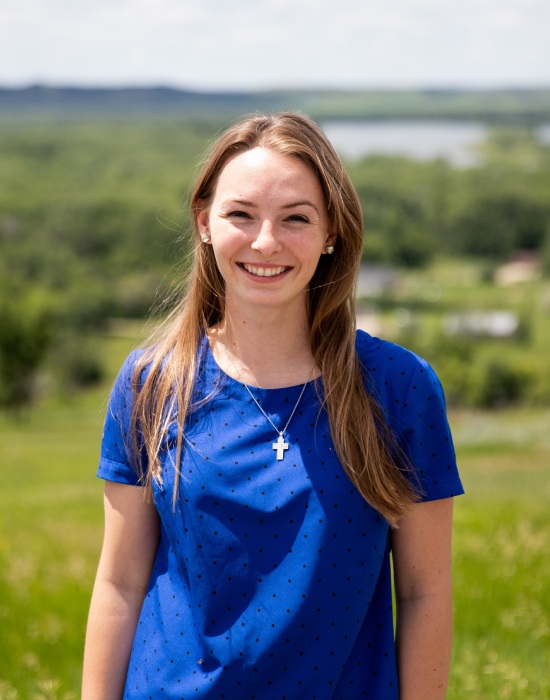 Young woman with long hair wearing a blue shirt and cross necklace standing outdoors with a scenic background of fields, trees, and a body of water, looking like someone who provides patient services.