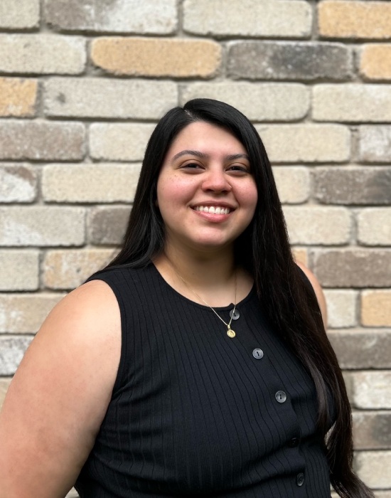 A woman with long dark hair, wearing a black sleeveless top and a necklace, is smiling in front of a brick wall, exuding the warmth and professionalism often found in Patient Services.