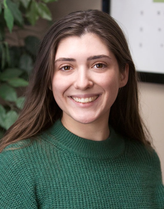 A young woman with long brown hair, wearing a green sweater, smiles warmly in front of a blurred background with green leaves. Her kind expression reflects her dedication to Patient Services.