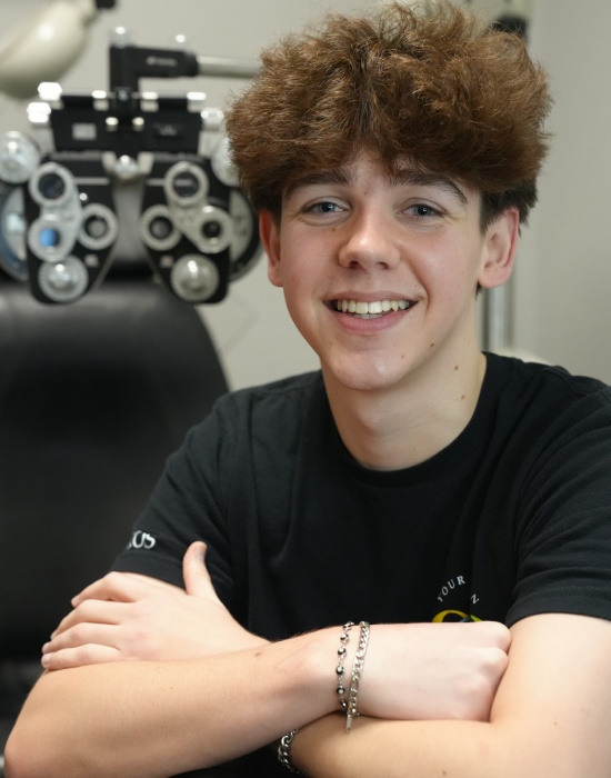 A young person with a broad smile sits in an optometrist's chair in Nampa, arms crossed, with an eye examination machine in the background.