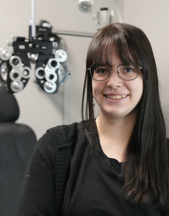 A smiling woman with glasses sitting in front of an ophthalmology phoropter at an eye doctor in Meridian.