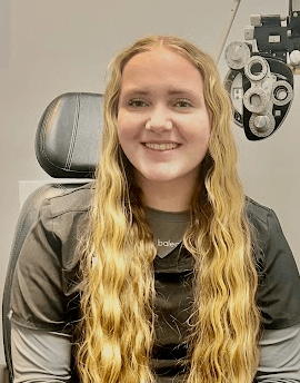 A person with long blonde hair is sitting in an eye examination chair, smiling at the camera, with optical equipment visible in the background. The friendly Eye Doctor Idaho Falls provides a welcoming atmosphere for all patients.