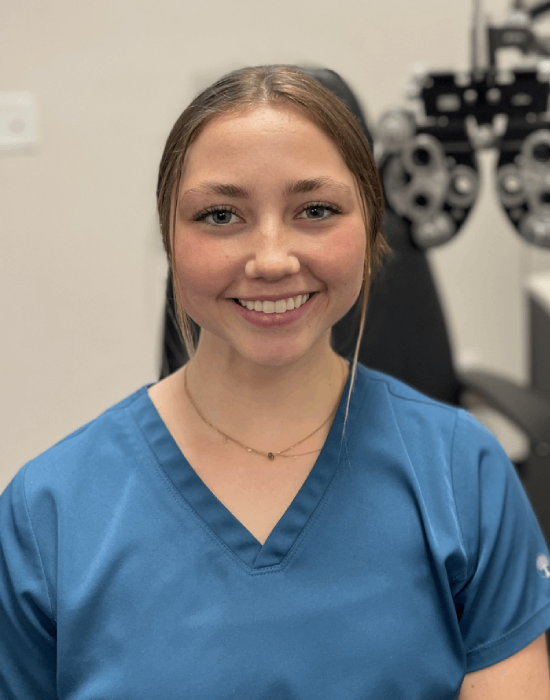 A woman with light brown hair in a low bun, wearing blue scrubs, smiles at the camera. She is standing in an Eye Doctor Pocatello office with diagnostic equipment in the background.