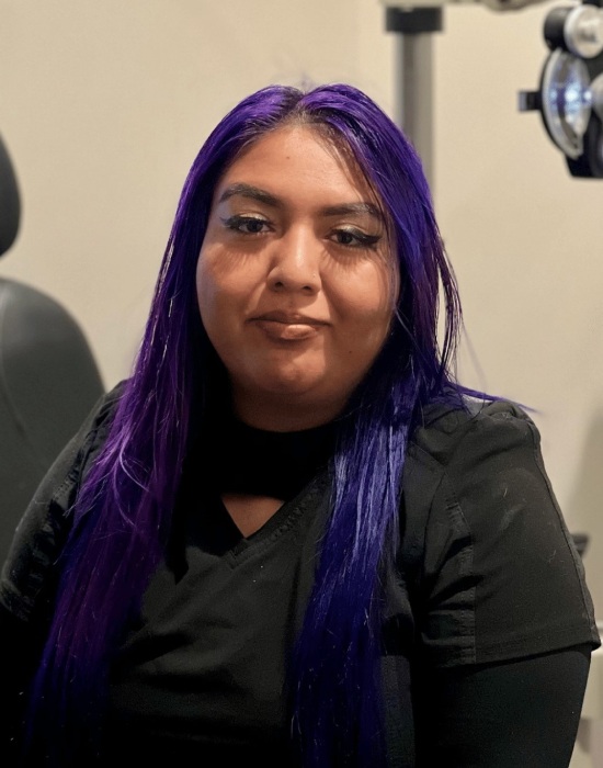 A person with long, straight, vibrant purple hair wearing a black shirt, seated and looking at the camera, could be waiting for an appointment with Eye Doctor Ogden.