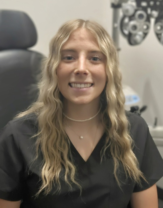 A woman with long, wavy hair and wearing a black blouse sits in front of an eye examination machine, smiling at the camera at Eye Doctor Ogden.