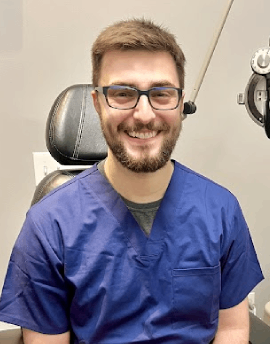 A man with glasses and a beard, wearing blue medical scrubs, smiles while sitting in an exam chair at a medical facility. This friendly Eye Doctor Idaho Falls ensures his patients receive the best care possible.