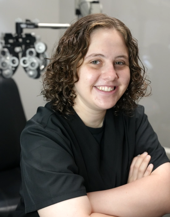 A Boise eye doctor with curly hair and wearing a black shirt smiles while sitting with arms crossed. An optometry device is in the background.