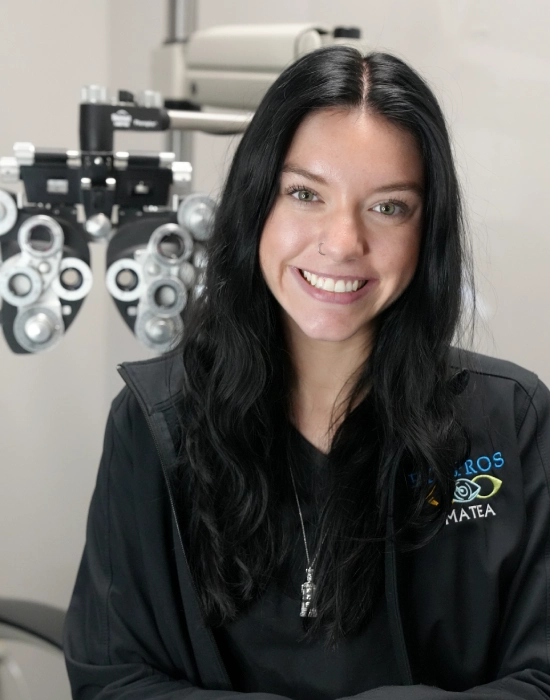 A person with long black hair and a nose ring is smiling and standing in front of an eye examination device, wearing a black jacket with "VRSOS" and "MATEA" embroidered on it, at their Boise eye doctor's office.