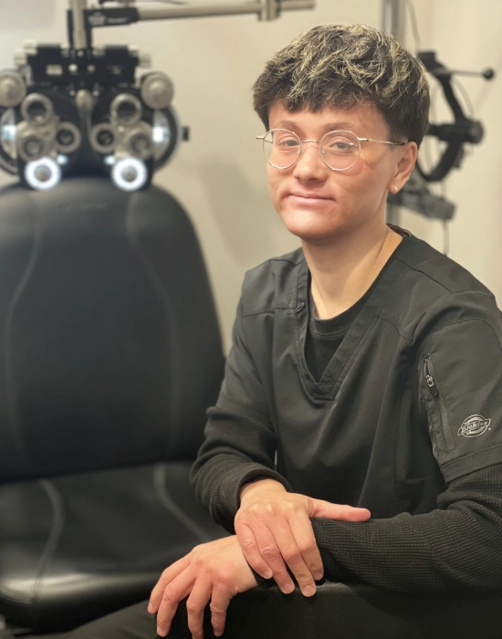 Person wearing glasses and black clothing sits in front of optometric equipment at the Layton eye doctor, smiling slightly, with hands resting on their lap.