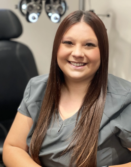 A woman with long brown hair is sitting and smiling at the camera. She is wearing a grey uniform, and an office chair and medical equipment are visible in the background, suggesting she might be a Layton eye doctor.