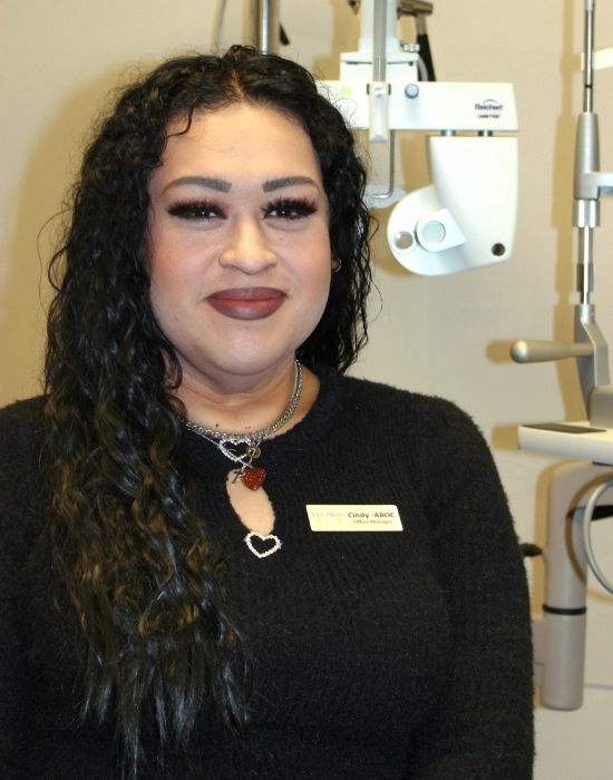 A Boise eye doctor with long curly hair, wearing a black sweater and nametag, stands confidently in front of ophthalmic equipment.