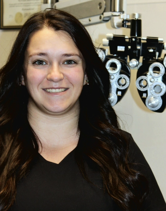 A smiling person with long dark hair stands in front of optometry equipment at a Boise eye doctor clinic.