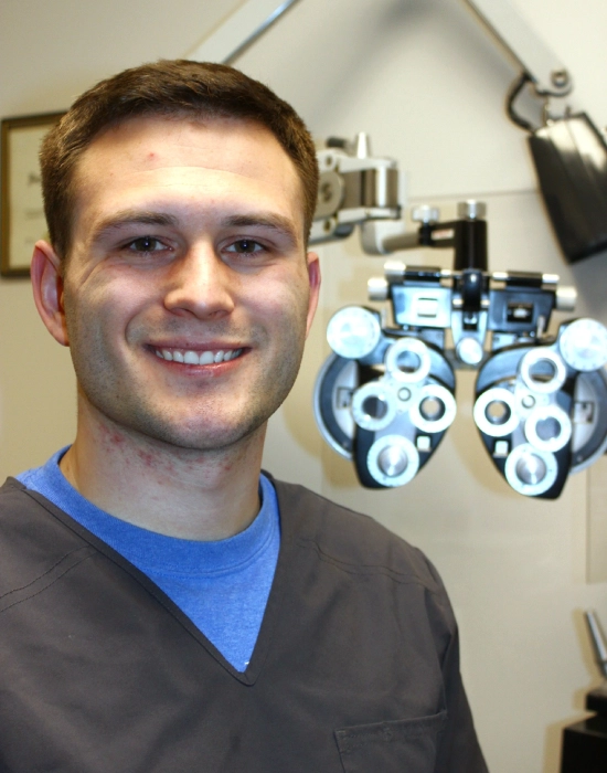 A smiling person stands proudly next to the optometry equipment in an exam room, embodying the professional dedication of a Boise eye doctor.