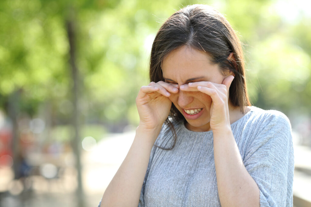 A woman in a gray sweater rubs her eyes outdoors on a sunny day, enjoying the vibrant surroundings. Maintaining healthy eyes is vital to prevent eye infections, especially when enjoying nature's beauty with a blurred green backdrop.