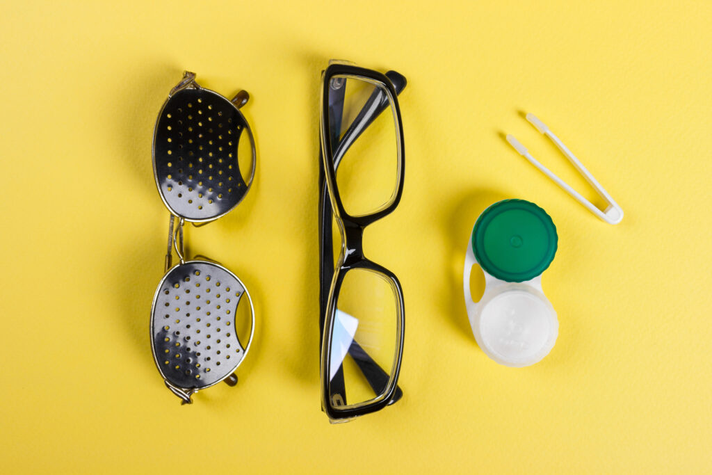 Flat lay of eyeglasses, pinhole glasses, and a contact lens case with tweezers on a vibrant yellow background, showcasing vision care essentials.