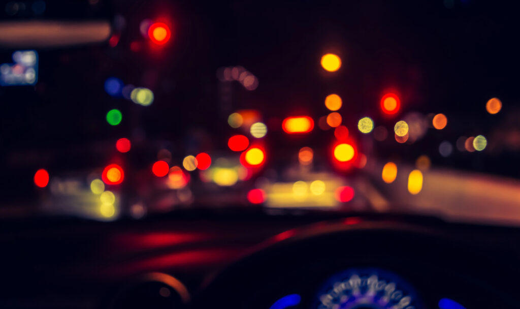 The blurred view from inside a vehicle at night, with unfocused traffic and car lights ahead, captures the challenge of night driving for those with astigmatism. A dashboard sits partially visible in the foreground, adding to this hazy yet familiar scene.