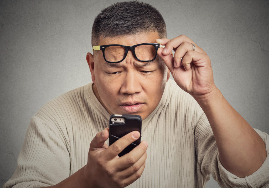 A man squints while holding his smartphone close, adjusting his glasses as he looks intently at the screen. Sporting a light-colored sweater against a plain background, he's focused on correcting vision issues, balancing monovision for both near and far vision tasks.