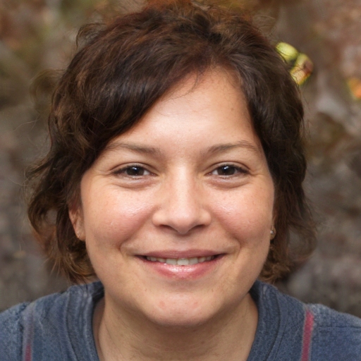 A person with short brown hair and a gray top is smiling at the camera in Idaho Falls, with a blurred outdoor background.
