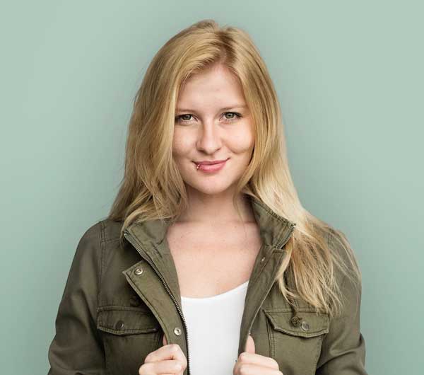 Woman with long blonde hair wearing a green jacket over a white top, standing in front of a plain light green background.