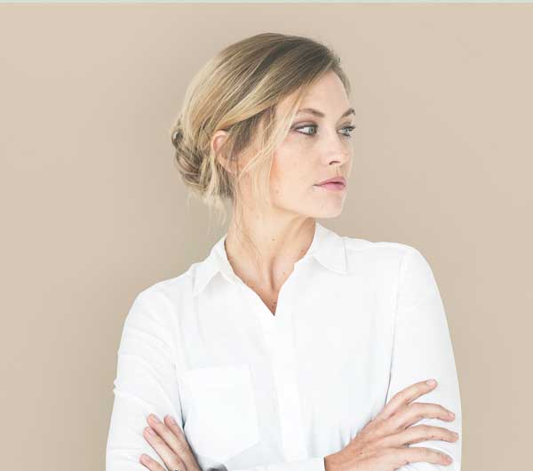 A woman with blonde hair in a low bun, wearing a white shirt, stands with arms crossed and looks to her left against a beige background.