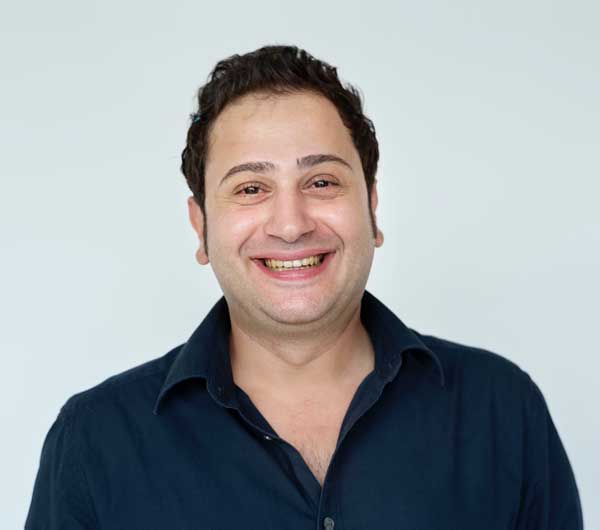 A smiling man with dark hair, wearing a dark blue collared shirt, poses against a plain light background.