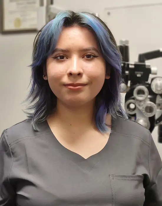 A person with short, blue-purple hair wearing gray scrubs stands in front of optometry equipment, ready to assist patients as a Spanish Fork eye doctor in a clinical setting.