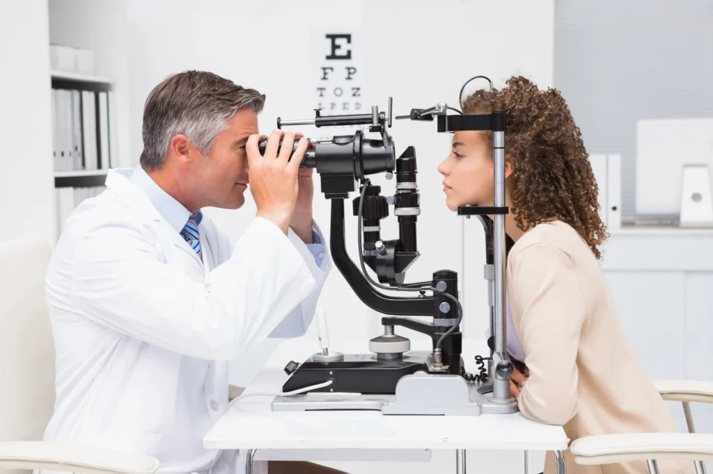 An eye doctor examines a woman’s eyes with a slit lamp in a clinical office, with an eye chart visible in the background—offering same-day eye exams in Coeur d’Alene.