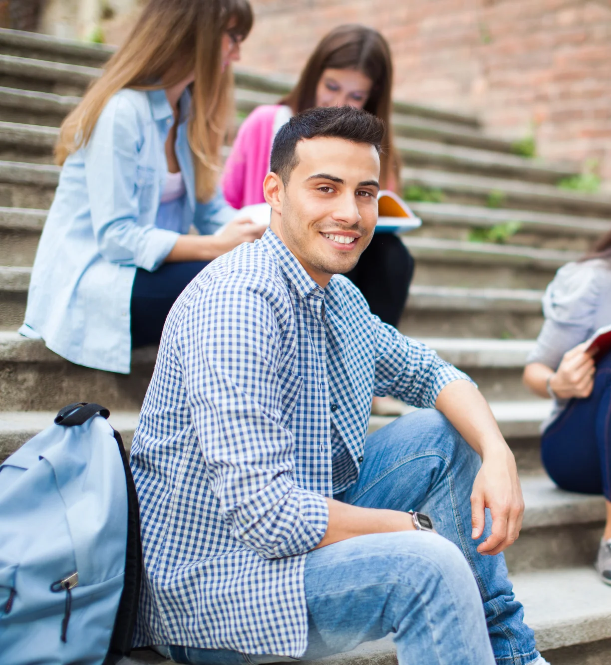 Young man sitting on outdoor steps smiling at the camera, with three other people behind him—fellow Weber State students engaged in conversation and reading.