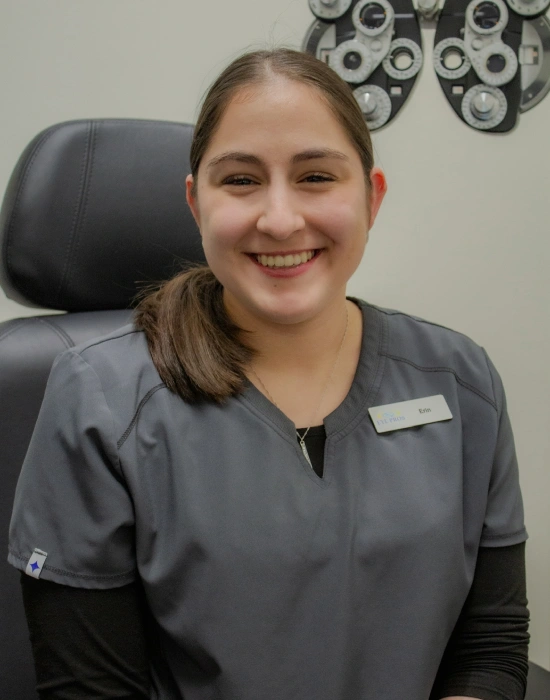 A woman wearing gray scrubs and a name tag sits in an eye exam room, smiling at the camera. Optical equipment is visible in the background, highlighting her role as a Twin Falls eye doctor.