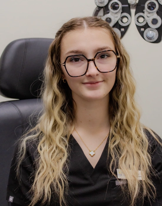 A woman with long wavy blonde hair and glasses sits in a Twin Falls eye doctor’s office, wearing black scrubs and a name tag. Optical equipment is visible in the background.